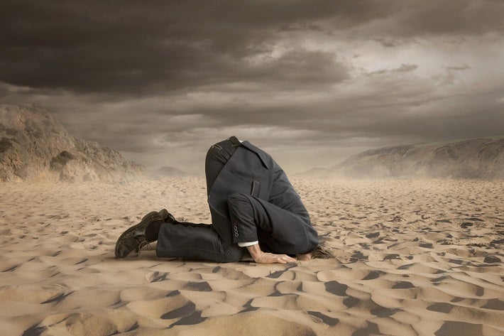 Image of business man on his knees with his head burried in the sand.